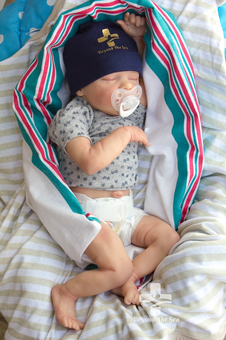 May include: A sleeping baby doll wearing a blue and gold hat with the text "Beyond the Sea" and a white pacifier. The doll is wrapped in a striped blanket with red, white, and teal stripes.