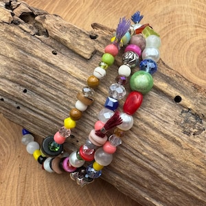 Könnte beinhalten: Drei Armbänder mit Perlen mit einer Vielzahl von farbigen Perlen, darunter Perlen, facettierte Kristalle und Holzperlen. Die Armbänder sind auf einem Stück verwittertem Holz angeordnet und zeigen eine Mischung aus Texturen und Farben.