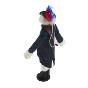 May include: A felted figure of a soldier in a dark blue coat with white trim, black boots, and a brown strap. The soldier's hat is adorned with red and blue feathers. A white string is attached to the top of the figure.