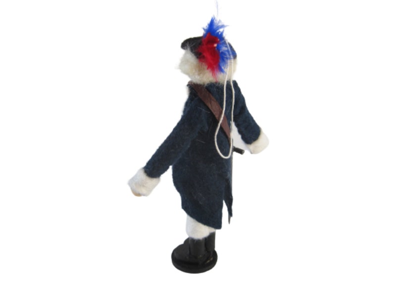 May include: A felted figure of a soldier in a dark blue coat with white trim, black boots, and a brown strap. The soldier's hat is adorned with red and blue feathers. A white string is attached to the top of the figure.
