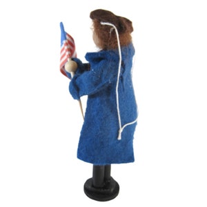 K&ouml;nnte beinhalten: Eine Filzpuppe, die einen blauen Mantel tr&auml;gt und eine kleine amerikanische Flagge h&auml;lt. Die Puppe steht auf einem Holzsockel.