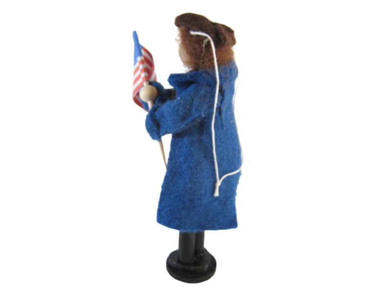 K&ouml;nnte beinhalten: Eine Filzpuppe, die einen blauen Mantel tr&auml;gt und eine kleine amerikanische Flagge h&auml;lt. Die Puppe steht auf einem Holzsockel.