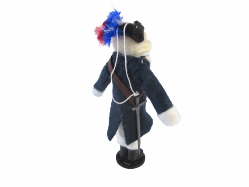 May include: A handmade doll dressed in a dark blue coat with white cuffs and a brown strap. The doll has a white face, black hat, and a decorative feather plume with red and blue accents. A small sword hangs at the doll's side.