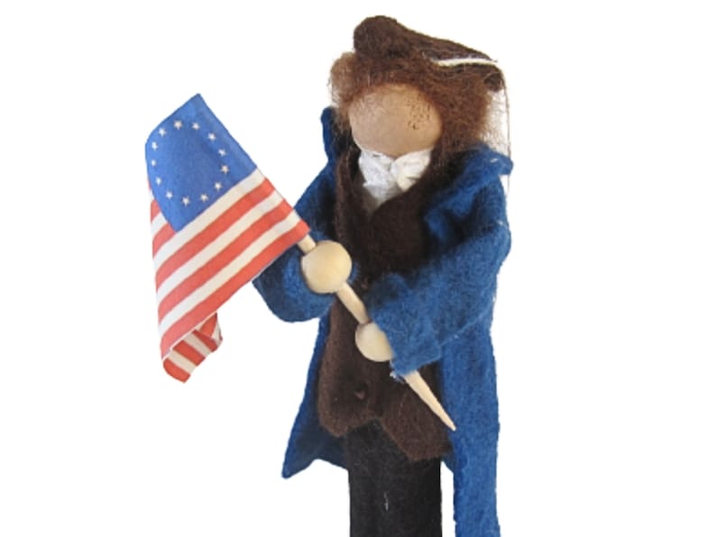 K&ouml;nnte beinhalten: Eine Filzpuppe, die einen blauen Mantel und eine braune Weste tr&auml;gt und eine kleine amerikanische Flagge mit 13 Sternen h&auml;lt.