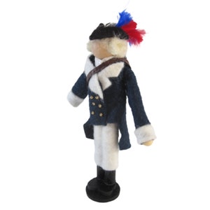 May include: A handcrafted doll dressed in a navy blue coat with white accents, gold buttons, and a tricorn hat adorned with red and blue feathers. The doll is wearing white pants and black boots.