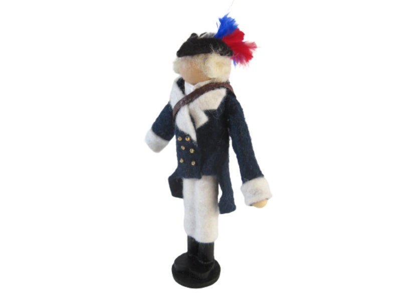 May include: A handcrafted doll dressed in a navy blue coat with white accents, gold buttons, and a tricorn hat adorned with red and blue feathers. The doll is wearing white pants and black boots.