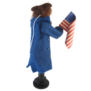 K&ouml;nnte beinhalten: Eine Filzpuppe in einem blauen Mantel und einem braunen Hut h&auml;lt eine kleine amerikanische Flagge mit 13 Sternen.