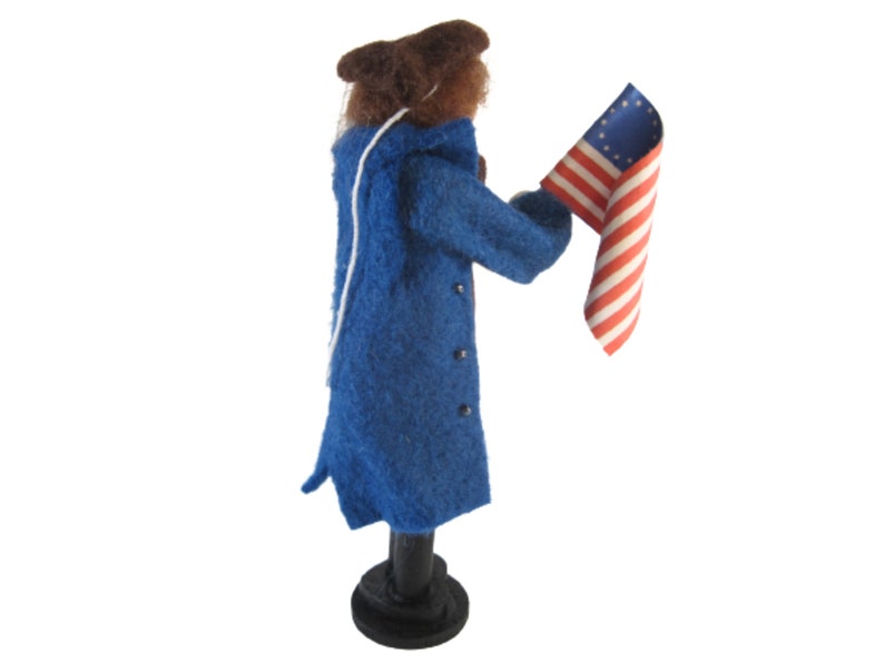 K&ouml;nnte beinhalten: Eine Filzpuppe in einem blauen Mantel und einem braunen Hut h&auml;lt eine kleine amerikanische Flagge mit 13 Sternen.