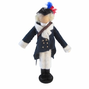 May include: A handcrafted doll dressed in a colonial-era military uniform. The doll features a dark blue coat with gold buttons, white pants, and a tricorn hat with red and blue accents. The doll stands on a black base.