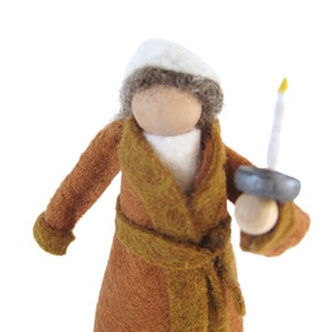 Puede incluir: Una muñeca de fieltro que lleva una túnica marrón con una capucha blanca y sostiene una vela blanca con una llama amarilla.