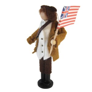 May include: A felt doll of a historical figure, wearing a brown coat, white waistcoat, and dark trousers. The doll holds a small flag with a red and white striped pattern and a blue field with white stars. The doll stands on a black base.