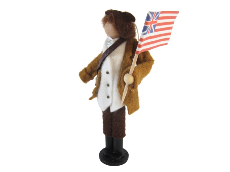 May include: A felt doll of a historical figure, wearing a brown coat, white waistcoat, and dark trousers. The doll holds a small flag with a red and white striped pattern and a blue field with white stars. The doll stands on a black base.