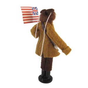 May include: A miniature figure of a person in a brown coat and hat, holding a flag with red and white stripes and a blue canton with white stars and a Union Jack. The figure is standing on a black base, with a white string attached to the flag.