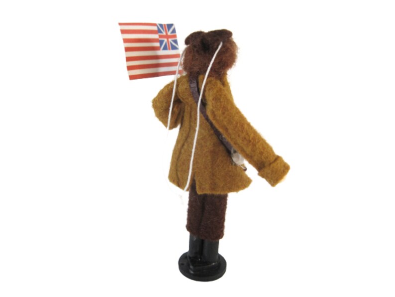 May include: A miniature figure of a person in a brown coat and hat, holding a flag with red and white stripes and a blue canton with white stars and a Union Jack. The figure is standing on a black base, with a white string attached to the flag.