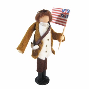 May include: A felted figure of a colonial soldier, dressed in a brown coat, white vest, and brown breeches. The figure holds a flag with red and white stripes and a blue canton with white stars. The figure stands on a black base.
