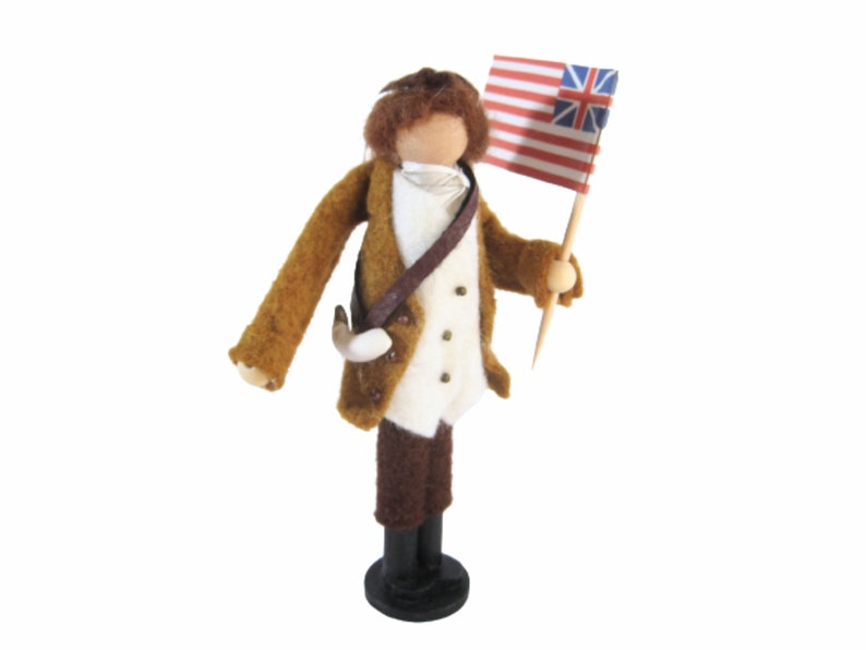 May include: A felted figure of a colonial soldier, dressed in a brown coat, white vest, and brown breeches. The figure holds a flag with red and white stripes and a blue canton with white stars. The figure stands on a black base.