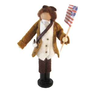 May include: A handmade doll depicting a soldier in a brown coat, white vest, and brown trousers. The doll holds a small flag with red and white stripes and blue and white symbols. The doll stands on a black base.