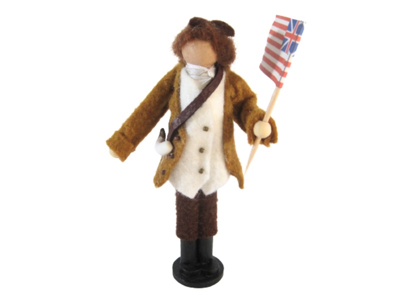 May include: A handmade doll depicting a soldier in a brown coat, white vest, and brown trousers. The doll holds a small flag with red and white stripes and blue and white symbols. The doll stands on a black base.
