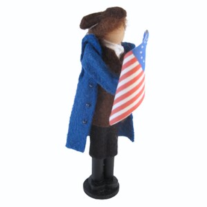 K&ouml;nnte beinhalten: Eine Filzpuppe, die einen blauen Mantel und eine braune Hose tr&auml;gt und eine amerikanische Flagge h&auml;lt. Die Puppe tr&auml;gt einen braunen Hut und hat ein braunes Gesicht.