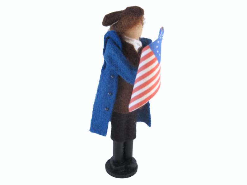 K&ouml;nnte beinhalten: Eine Filzpuppe, die einen blauen Mantel und eine braune Hose tr&auml;gt und eine amerikanische Flagge h&auml;lt. Die Puppe tr&auml;gt einen braunen Hut und hat ein braunes Gesicht.