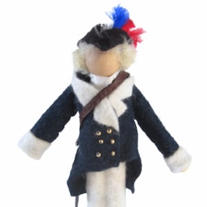 May include: A handcrafted doll dressed in a navy blue coat with gold buttons, white cuffs, and a white collar. The doll has a black hat with a red, white, and blue plume, and a brown strap across the chest. The doll is made of felt.