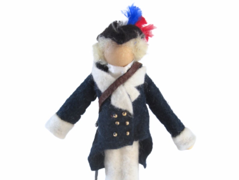 May include: A handcrafted doll dressed in a navy blue coat with gold buttons, white cuffs, and a white collar. The doll has a black hat with a red, white, and blue plume, and a brown strap across the chest. The doll is made of felt.