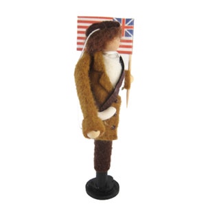 May include: A felt doll of a soldier holding a flag with red and white stripes and a Union Jack. The doll is dressed in a brown coat, dark trousers, and a white shirt. It stands on a black base.