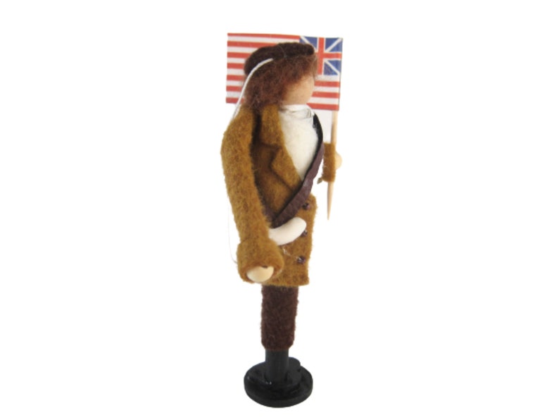 May include: A felt doll of a soldier holding a flag with red and white stripes and a Union Jack. The doll is dressed in a brown coat, dark trousers, and a white shirt. It stands on a black base.