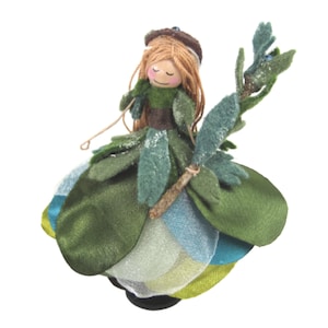 May include: A whimsical felt doll with a green and blue dress, brown hair, and a twig in her hand. The doll is wearing a brown felt hat and has a painted face.