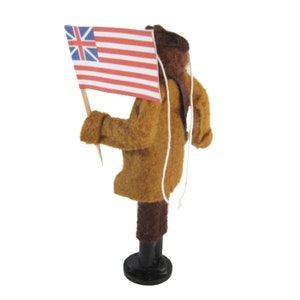 May include: A felt figure of a person in a brown coat and dark brown trousers, holding a small flag with the British Union Jack and red and white stripes. The figure stands on a black base, with a white string hanging down.