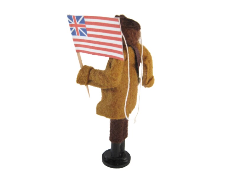 May include: A felt figure of a person in a brown coat and dark brown trousers, holding a small flag with the British Union Jack and red and white stripes. The figure stands on a black base, with a white string hanging down.