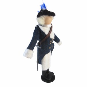 May include: A handcrafted felt doll depicting a soldier in a dark blue coat with white trim and gold buttons. The doll has a black hat with a blue plume, a brown strap, and a sword. The doll is standing on a black base.