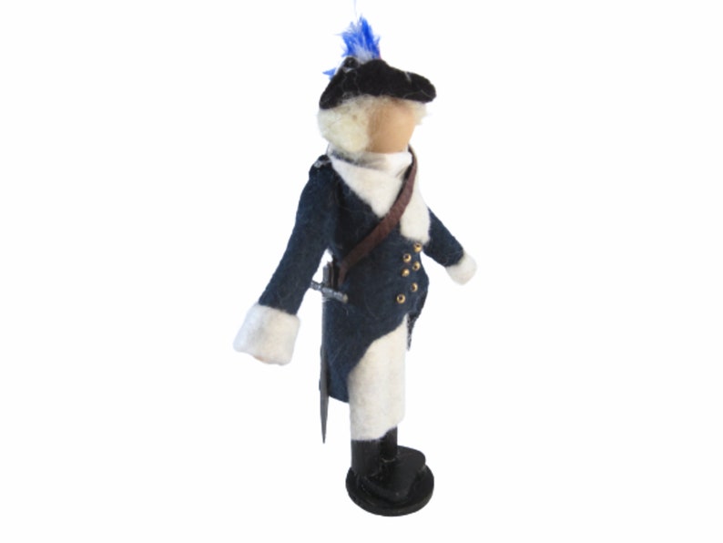 May include: A handcrafted felt doll depicting a soldier in a dark blue coat with white trim and gold buttons. The doll has a black hat with a blue plume, a brown strap, and a sword. The doll is standing on a black base.