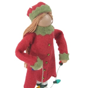 May include: A handmade Christmas elf doll wearing a red felt coat, hat, and collar with green trim. The doll has blonde yarn hair and is holding a string of miniature Christmas lights. The elf has black and white striped legs.