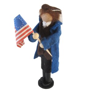 K&ouml;nnte beinhalten: Eine Filzpuppe, die einen blauen Mantel und eine braune Hose tr&auml;gt, h&auml;lt eine kleine amerikanische Flagge mit 13 Sternen.