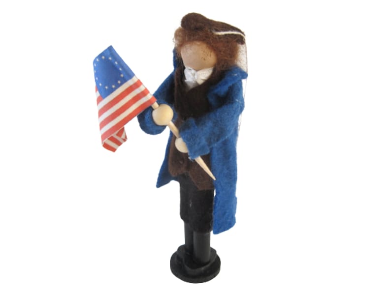 K&ouml;nnte beinhalten: Eine Filzpuppe, die einen blauen Mantel und eine braune Hose tr&auml;gt, h&auml;lt eine kleine amerikanische Flagge mit 13 Sternen.