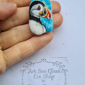 Puede incluir: Pintura de roca pintada a mano de un frailecillo con cabeza negra, cuerpo blanco y pico naranja sobre un fondo azul. La roca está pintada con un cielo azul claro y nubes blancas. La roca está sostenida en una mano. La imagen incluye el texto "Art Sea Glass Eco Shop by Dorota Borowieckaa".