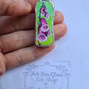 Peut inclure: Un pendentif fait à la main représentant une peinture de digitales pourpres sur un fond vert. L'œuvre est encadrée dans un cadre en métal argenté. Le pendentif est tenu dans une main, avec le texte "Art Sea Glass Eco Shop by Dorota Borowiecka" visible.