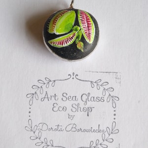 May include: A round glass pendant with a black background and a painted image of a green and red flower. The pendant is attached to a silver chain. The pendant is labeled "Art Sea Glass Eco Shop by Dorota Borowiecka".