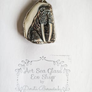 Puede incluir: Un colgante de plata con una imagen pintada a mano de una morsa sobre un fondo blanco. El colgante está hecho de vidrio de mar y forma parte de la tienda ecológica Art Sea Glass de Dorota Borowiecka.