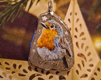 May include: A hand-painted pendant featuring a robin bird with orange breast and blue wings. The pendant is set in a silver-colored metal frame and has a loop for hanging. The background includes a decorative star and evergreen branches.