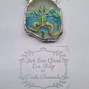 Peut inclure: Une grenouille verte peinte sur un morceau de verre de mer, encadrée d'argent, avec une bélière en argent pour la suspension. Le verre est entouré de peinture bleue pour représenter l'eau. La pièce est signée "Art Sea Glass Eco Shop by Dorota Borowicka".
