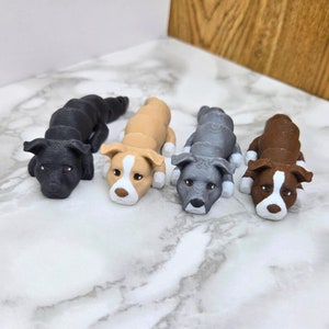 Peut inclure: Quatre petites figurines de chiens peintes à la main dans différentes couleurs : noir, marron, beige et gris. Les chiens sont tous en position recroquevillée, la tête reposant sur leurs pattes.