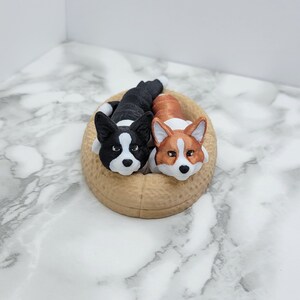 May include: Two plush corgi dogs, one black and white and one brown and white, are lying in a tan-colored dog bed.