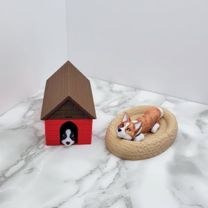 May include: A brown and red dog house with a black and white dog peeking out of the door. A brown and white dog is lying in a brown bed.