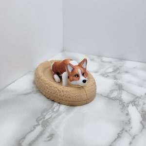 May include: A brown and white dog figurine laying in a tan, oval-shaped bed.