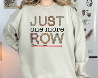 May include: A light beige sweatshirt with the text "JUST one more ROW" in a crocheted font. The letters are in shades of pink, beige, and green. A pink crochet hook is below the text.