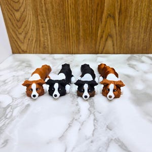 Könnte beinhalten: Vier Miniatur-Border-Collie-Hunde-Figuren, die auf dem Bauch liegen. Die Hunde sind braun und schwarz mit weißen Markierungen. Die Figuren sind aus glattem, bemaltem Material.