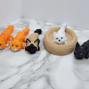 Op de afbeelding: Vier kleine kattenbeeldjes, twee oranje, &eacute;&eacute;n zwart en &eacute;&eacute;n wit. De witte kat ligt in een bruin kattenbed.