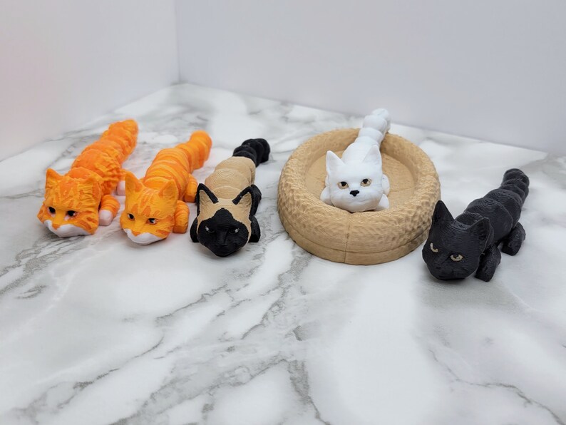 Op de afbeelding: Vier kleine kattenbeeldjes, twee oranje, &eacute;&eacute;n zwart en &eacute;&eacute;n wit. De witte kat ligt in een bruin kattenbed.